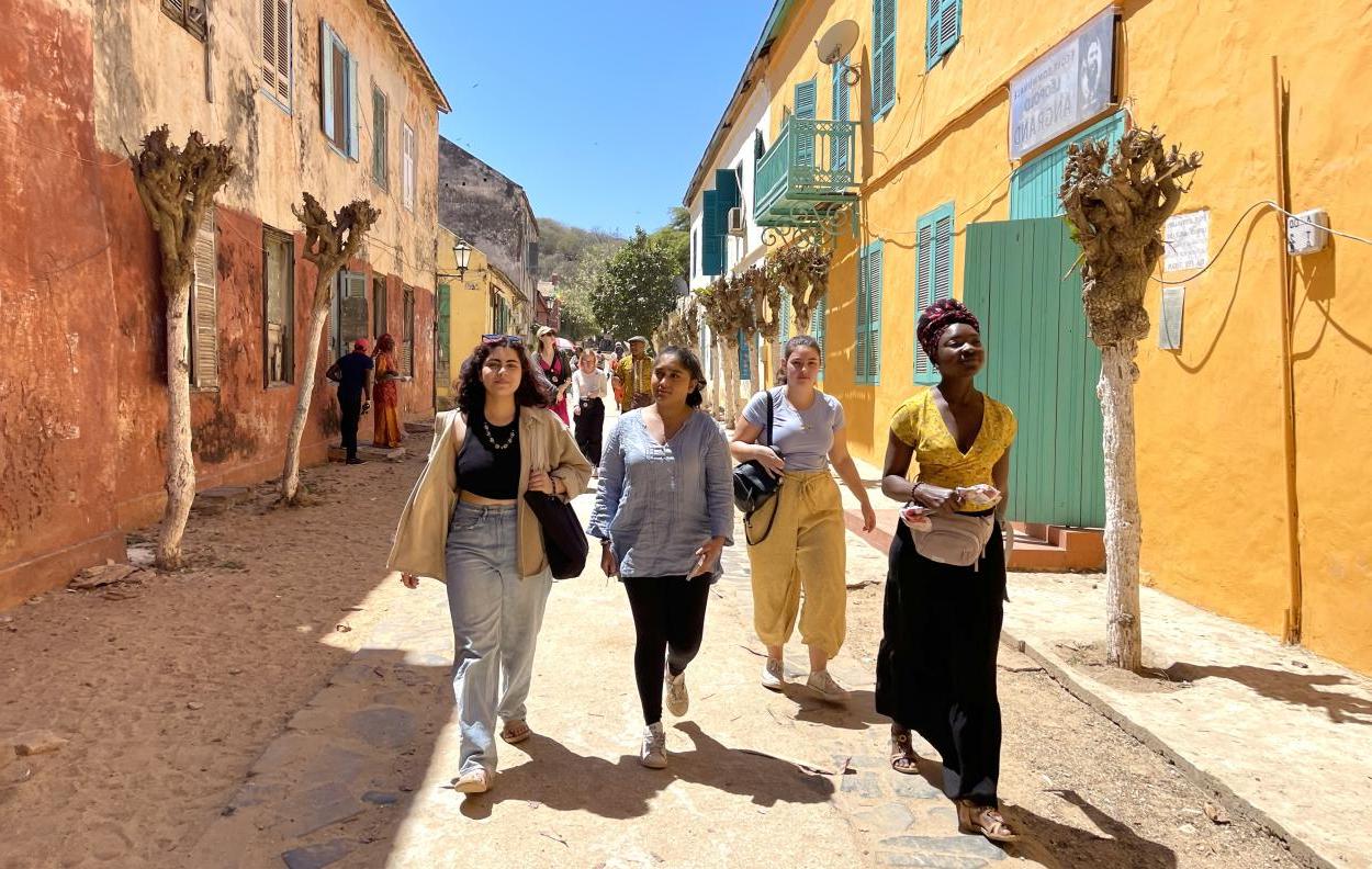 Four students walk down a dirt road with brightly colored two-storey buildings on either side.
